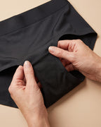 Mani che aprono lo slip LEIDRY™ Giorno nero e mostrano la zona assorbente interna